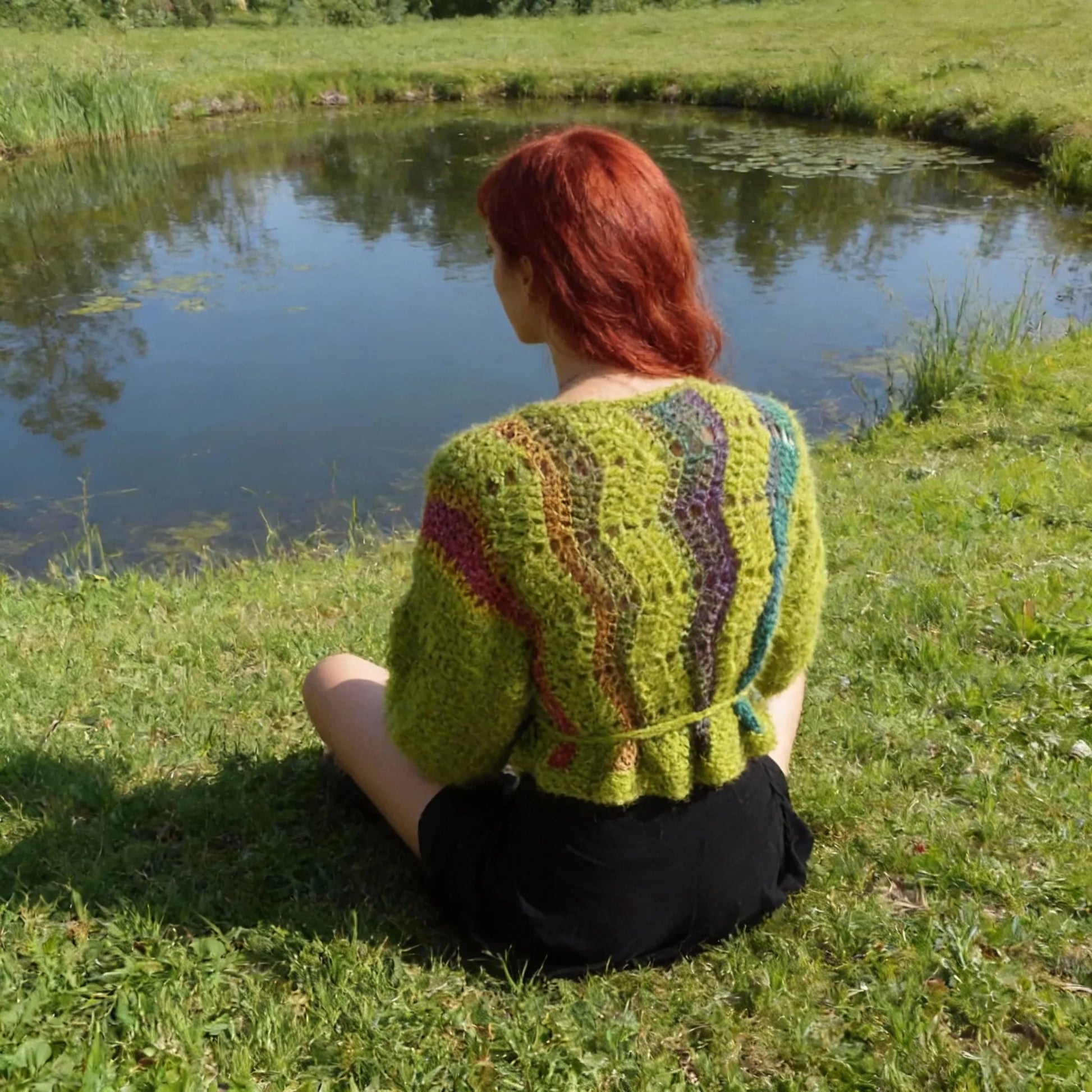 Gilet au crochet Avocado de Cachalabibi -vert anis avec rayures verticales multicolores - vue de dos assise au bord d'un étang