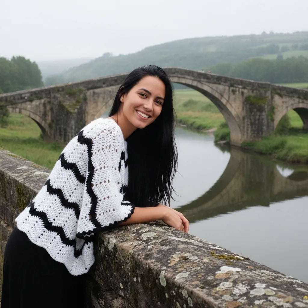 Poncho au crochet Bianconero de Cachalabibi - vue de face sur un pont en pierre avec rivière et collines verdoyantes